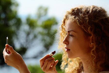 Pretty cute woman or sexy girl with curly blond hair put red lipstick on lips with mirror on green natural background sunny outdoor on balcony on summer dayの写真素材