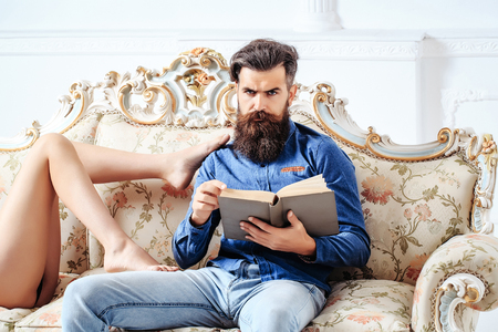 Handsome bearded man in blue shirt and denim with book and bare female legs touch him on luxurious baroque couch on white backgroundの写真素材