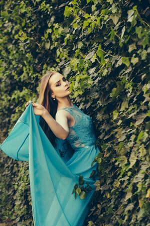pretty young sexy woman or girl with cute face and long hair in fashionable blue dress sunny outdoor on natural green leaves wall backgroundの写真素材