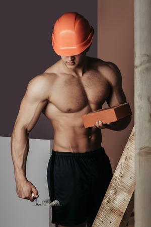 young handsome macho man builder with sexy muscular athletic strong body has bare torso and strong belly with six packs or abs in orange hard hat or helmet holds brick on wood ladderの写真素材