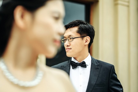Chinese groom handsome man asian young male in eyeglasses and elegant black suit with bow tie stands outdoors on pretty bride silhouetteの写真素材
