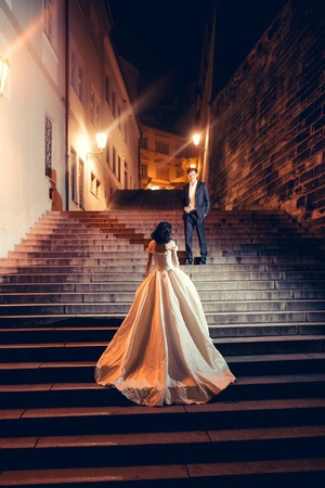Pretty girl bride young chinese cute sexy woman in long white wedding dress stands on stone steps outdoors on city night lightsの写真素材