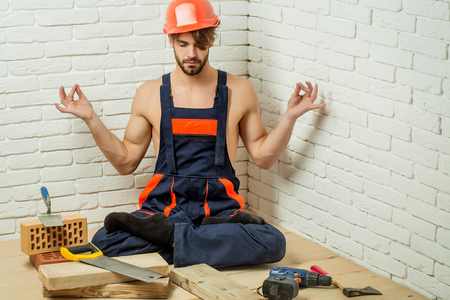 young handsome bearded macho man builder with sexy muscular athletic strong body has strong hands in blue uniform and orange hard hat or helmet with wood axe drill and saw on white brick wallの写真素材