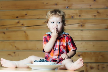 Cute baby boy child with blond curly hair eats delicious cake with blueberries sitting on wooden tableの写真素材