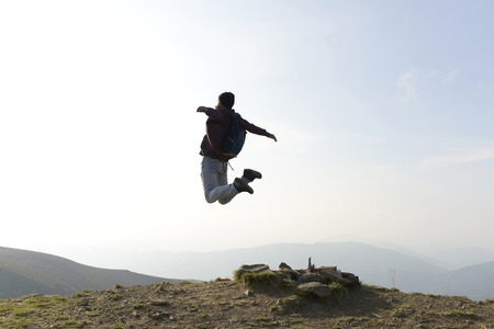 Handsome man hipster traveler with beard and moustache with backpack jumps on mountain top on foggy skyの写真素材