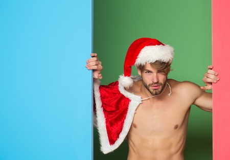 young handsome bearded sexy macho christmas man with long beard in red santa claus hat and coat with muscular bare torso in xmas holiday or new year on colorful background, copy spaceの写真素材
