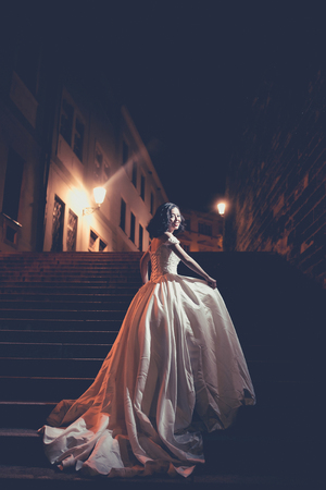Pretty girl bride young chinese cute sexy woman in long white wedding dress stands on stone steps outdoors on city night lightsの写真素材