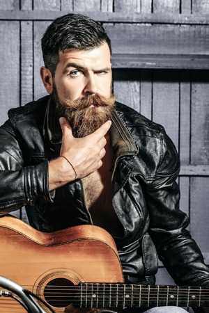 Frown bearded man hipster biker brutal male with beard and moustache in leather jacket sits on motorcycle with guitar on wooden backgroundの写真素材