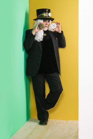 Senior man bearded male or watchmaker with blue lenses in eyes and white beard hair in black hat with mechanical metallic gears and cogwheels stands with clock on colorful wallの写真素材