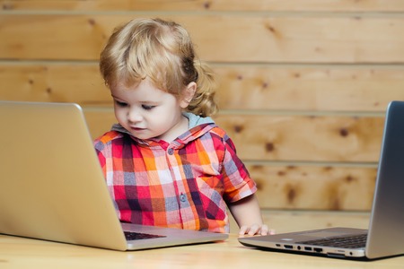 Cute baby boy child with blond curly hair plays on two laptop computers on wooden background