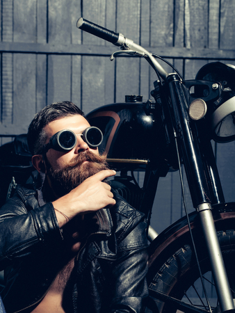 Bearded man hipster biker brutal male with beard and moustache in vintage sunglasses and leather jacket sits near motorcycle on wooden backgroundの写真素材