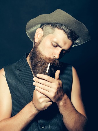 Bearded man with beard and moustache stylish hipster male in hat lights cigarette to smoke on grey backgroundの写真素材