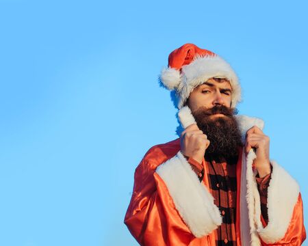 young handsome surprised bearded bad santa claus man with long beard in checkered shirt and red new year hat in christmas or xmas coat outdoor on blue sky background, copy spaceの写真素材