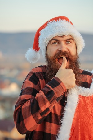 young handsome happy emotional bearded bad santa claus man with long beard in checkered shirt and red new year hat in christmas or xmas coat outdoor on blue sky background with thumb upの写真素材