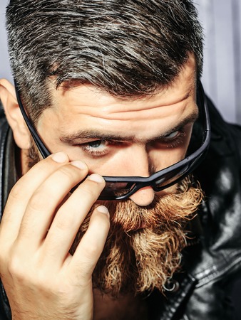 Frown bearded man hipster brutal male with beard and moustache in leather jacket looks above sunglasses on wooden backgroundの写真素材