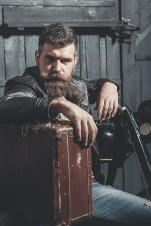 Bearded man hipster biker brutal male with beard and moustache in leather jacket sits near motorcycle with vintage suitcase on wooden backgroundの写真素材
