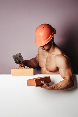 young handsome macho man builder with sexy muscular athletic strong body has bare torso and strong belly with six packs or abs in orange hard hat or helmet holds brick, copy spaceの写真素材