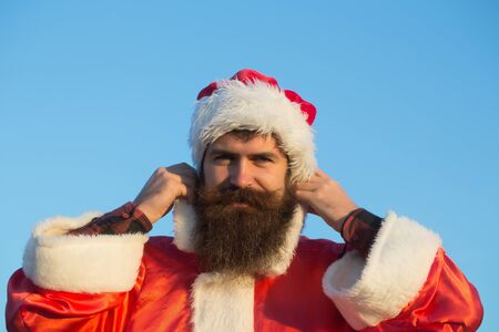 young handsome smiling bearded bad santa claus man with long beard in red new year hat in christmas or xmas coat sunny outdoor on blue sky backgroundの写真素材