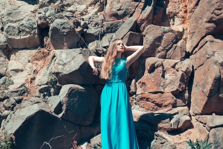 pretty young sexy woman or girl with cute face and long hair in fashionable blue dress sunny outdoor on stony wall backgroundの写真素材