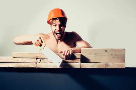 young handsome bearded macho man screaming builder with sexy muscular athletic strong body has strong hands in orange hard hat or helmet holds saw cutting wooden planks, copy spaceの写真素材