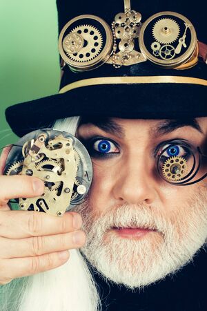 Senior man bearded male or watchmaker with blue lenses in eyes and white beard hair in black hat with mechanical metallic cogwheels keeps gears on green wallの写真素材