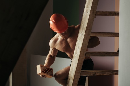 young handsome macho man builder with sexy muscular athletic strong body has bare torso and strong belly with six packs or abs in orange hard hat or helmet holds brick on wood ladderの写真素材