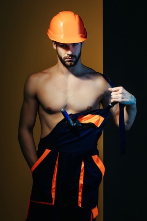 young handsome bearded macho man builder with sexy muscular athletic strong body has strong hands in blue uniform and orange hard hat or helmet with spatula in pocketの写真素材