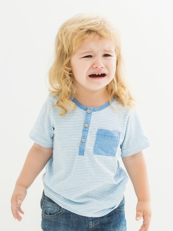 Cute baby boy child with curly blond hair in striped shirt and jeans cries isolated on white...