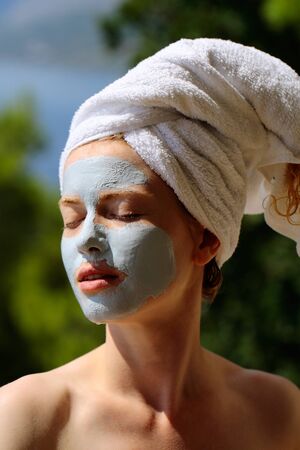 Pretty cute woman or girl with facial mask and white towel on head on sunny natural green backgroundの写真素材