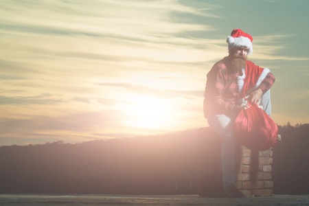 young handsome bearded bad santa claus man with long beard in checkered shirt jeans and red new year hat in christmas or xmas coat with toy sack on brick chimney in sunset, copy spaceの写真素材