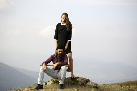 Couple of handsome bearded hipster in checkered shirt and pretty cute sexy young fashion girl or woman sitting on stone with ax on mountain top on natural backgroundの写真素材