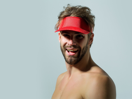 Joyful handsome man sexy young bearded male model with beard in red cap peak visor topless with nude torso and sexi body smiles on grey backgroundの写真素材