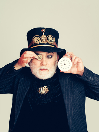 Senior bearded man or watchmaker with white beard in black hat with mechanical metallic gears keeps cogwheel and clock on grey wallの写真素材