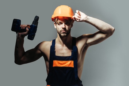 Handsome man builder repairman craftsman foreman or construction worker in orange hard hat and overall with drill on grey backgroundの写真素材