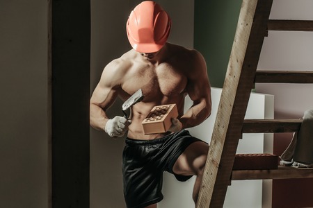 young handsome macho man builder with sexy muscular athletic strong body has bare torso and strong belly with six packs or abs in orange hard hat or helmet holds brick and tool near wood ladderの写真素材