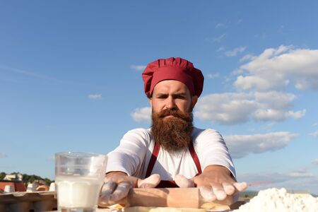 Man cook chef with beard on handsome face in white and red uniform cooking dough sunny outdoor on blue sky backgroundの写真素材