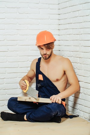 young handsome bearded macho man builder with sexy muscular athletic strong body has strong hands in blue uniform and orange hard hat or helmet holds wood and saw on white brick wall backgroundの写真素材