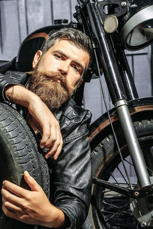 Bearded man hipster biker brutal male with beard and moustache in leather jacket sits on floor near motorcycle with auto wheel tire or tyre on wooden backgroundの写真素材