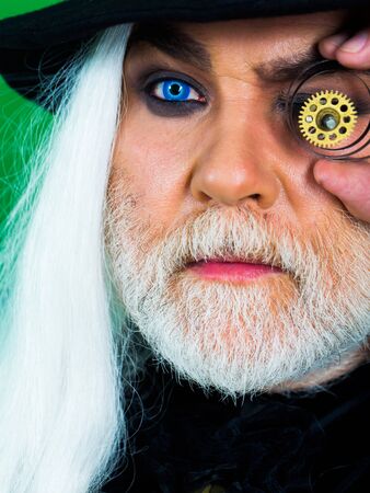 Senior bearded man or watchmaker with white beard and hair in black hat with watch mechanical metallic gears looks through cogwheel on green wallの写真素材