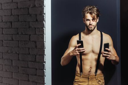 Handsome man confident fashion sexy young blond bearded male model with suspenders on topless sexi nude muscular torso holds two smartphones or mobile phones on grey backgroundの写真素材