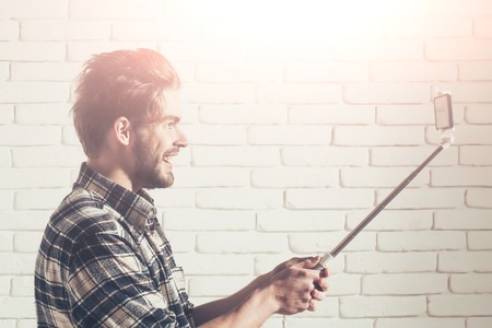Joyful handsome man young blond bearded male model with beard in plaid shirt uses smartphone or mobile phone on selfie stick on white brick backgroundの写真素材