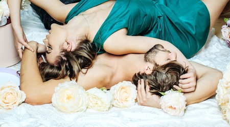 Sexy couple young beautiful cute lovers in love pretty girl and sexi man lying on backs on white bedsheet among flowers on floorの写真素材