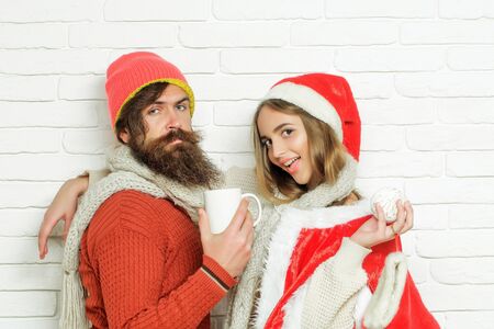 Young sensual Christmas couple of bearded man with gift box and pretty sexy girl or beautiful woman in red santa claus costume with xmas bauble and sock on white brick wallの写真素材