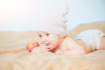 Little adorable happy hungry baby boy in white knitted hat with pompon on top with red ripe apple on beige blanketの写真素材