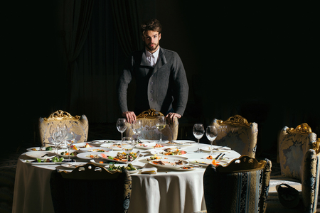 Handsome young man with beard and blond hair stands over table with leftovers or residues food on dirty plates after banquet dinner in restaurant on dark backgroundの写真素材