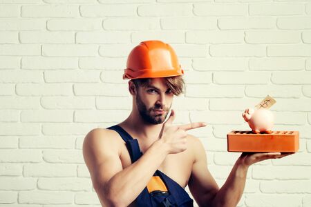 young handsome bearded macho man builder with sexy muscular athletic strong body has strong hands in orange hard hat or helmet holds pig dollar moneybox and brick on white wall backgroundの写真素材