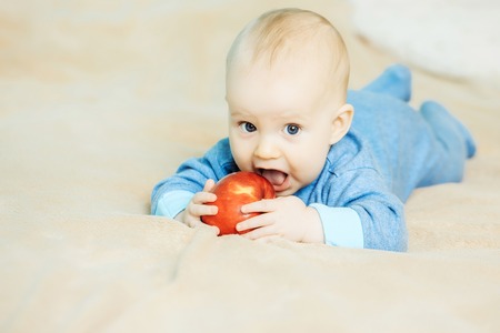 Little adorable baby boy in blue romper bites big red ripe apple on beige blanket