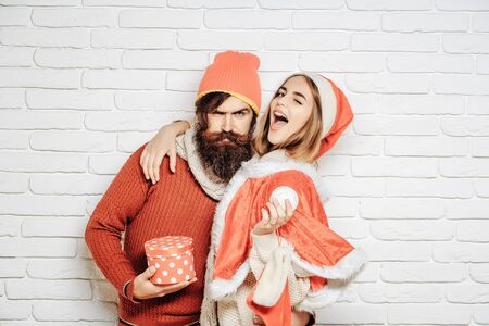 Young happy Christmas or new year couple of bearded man with gift box and pretty sexy girl or beautiful woman in red santa claus costume with xmas bauble and sock on white brick wallの写真素材