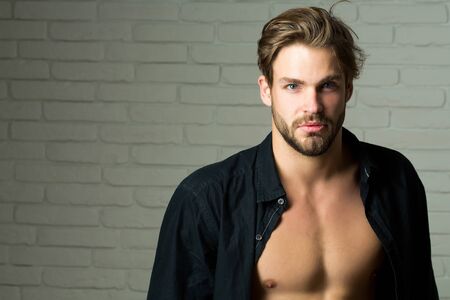 Sexy young handsome bearded man or guy with bare muscular chest in black shirt on white brick wall...