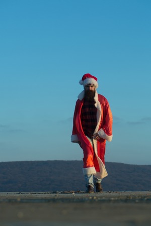 young handsome bearded bad santa claus man with long beard in checkered shirt jeans and red new year hat in christmas or xmas coat outdoor on blue sky background, copy spaceの写真素材